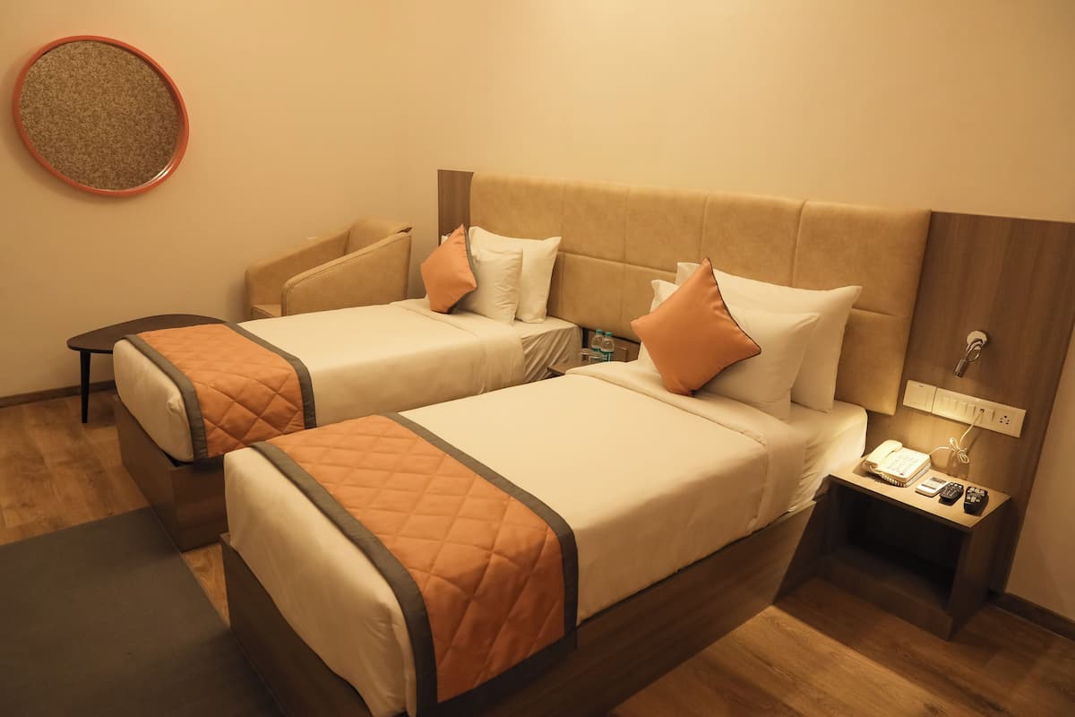 Quarto de hotel com duas camas, cada uma com um cobertor laranja acolchoado e almofadas brancas. Há uma pequena mesa de cabeceira com um telefone, controles remotos e uma garrafa.