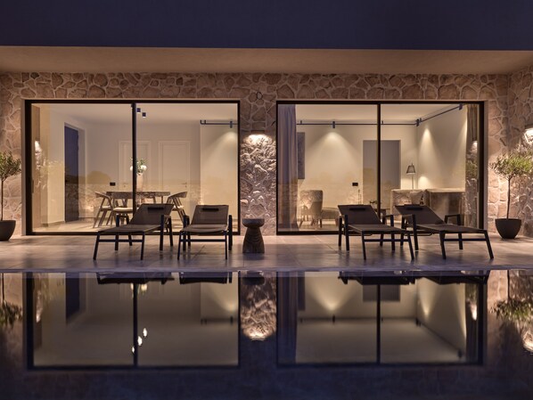 Un espace piscine extérieur moderne, doté de chaises longues et d’une table à manger, entouré de murs en pierre et de portes vitrées donnant sur un patio.