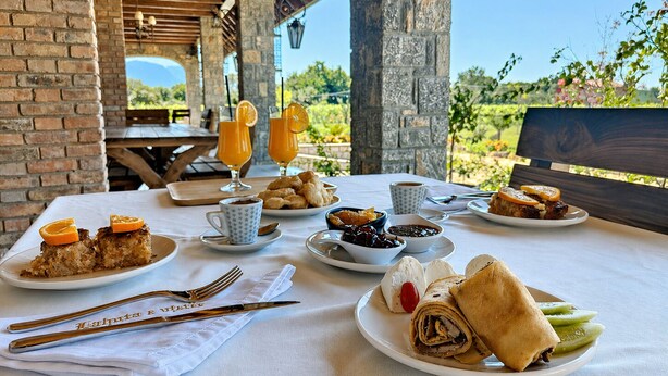 Une table de petit-déjeuner dressée avec des assiettes remplies de nourriture, des boissons et des couverts, avec en arrière-plan une vue sur la verdure et un mur de briques.