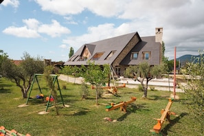 Una casa con techo inclinado, chimenea y jardín con columpios y sube y bajas.