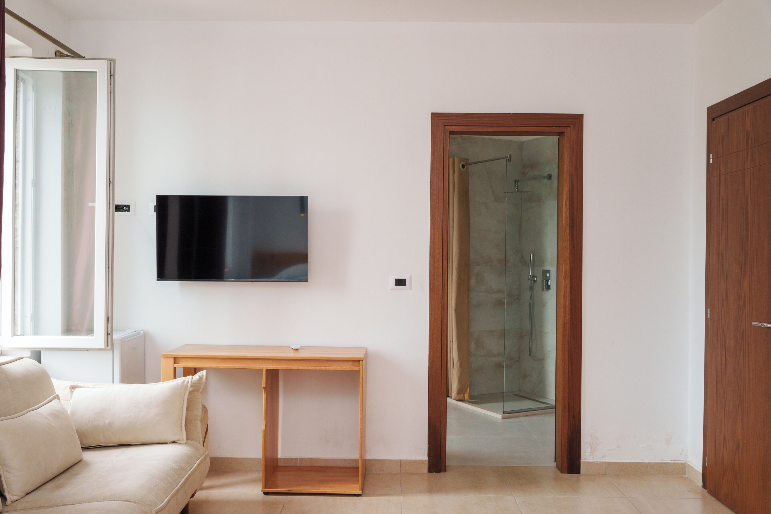 A room with a sofa, a wooden table, a TV mounted on the wall, and a glass shower door.