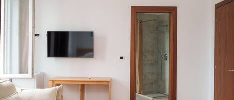 A room with a sofa, a wooden table, a TV mounted on the wall, and a glass shower door.