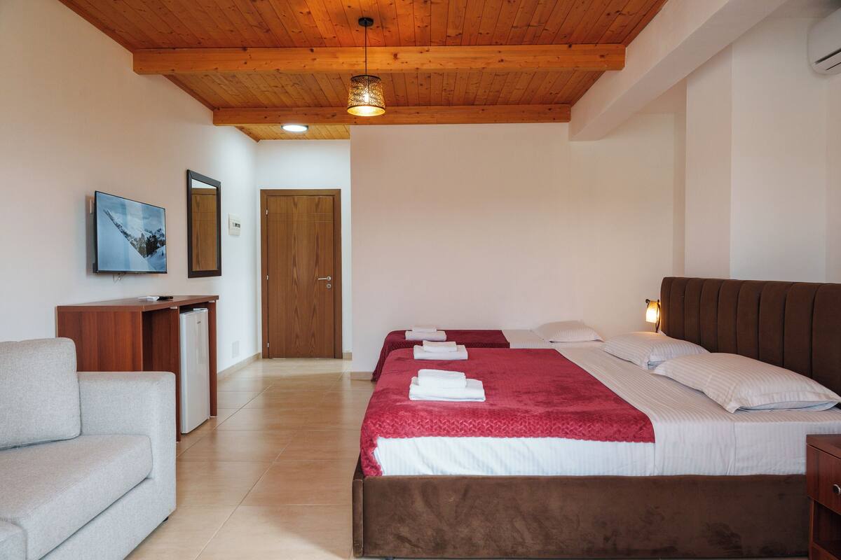 Habitació d'hotel amb sostre de fusta, llit amb roba de llit blanca i vermella, una petita taula de treball amb nevera i un sofà.