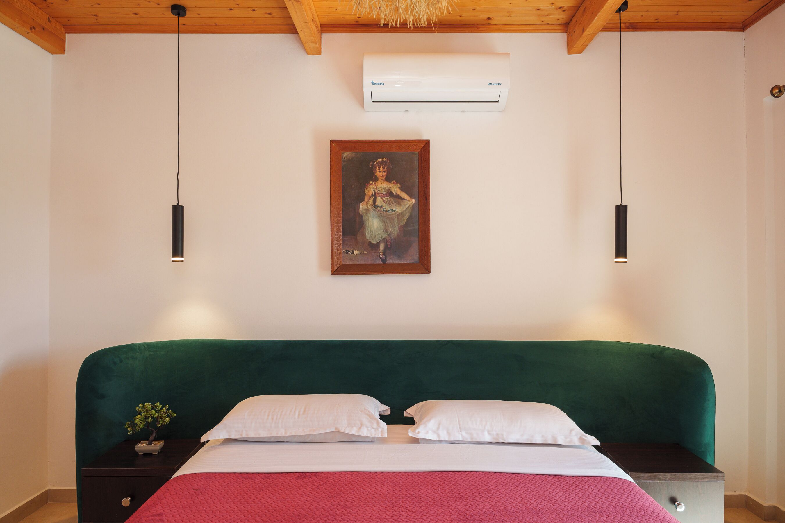A bedroom with a green upholstered headboard, a white bedspread, two white pillows, a nightstand with a small plant, and a painting on the wall.