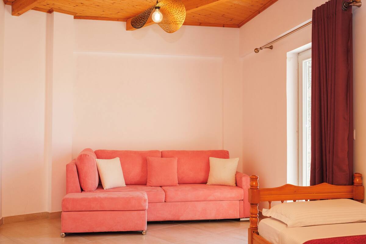 Habitació amb sostre de fusta, un sofà rosa amb coixins, estructura de llit de fusta i una finestra amb cortines.