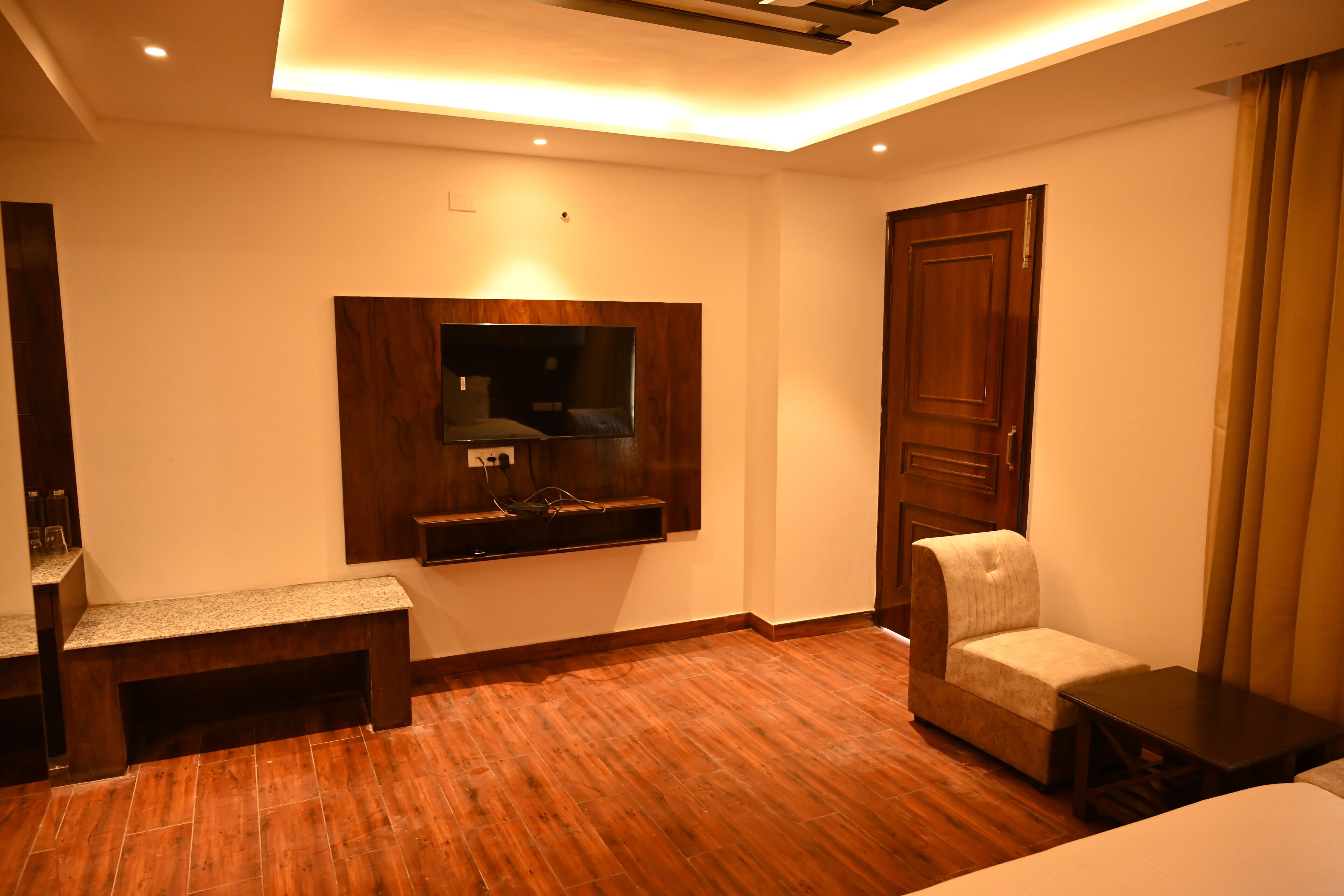 Habitación de hotel con piso de madera, un televisor montado en la pared, un banco, una silla y una puerta.
