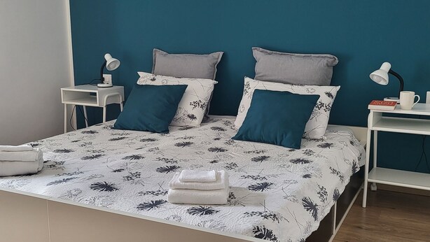 A neatly made bed with floral bedding, two bedside tables with lamps, and a small shelf with a book and mug. - Vrbo Property (Gorizia)