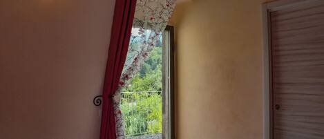 A bedroom with a bed, a window with a view of greenery, a wall-mounted light, and a door.