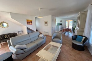 Una sala de estar con sofá, sillón y mesa de centro. Una escalera conduce a una zona de cocina con mesa y sillas de comedor.