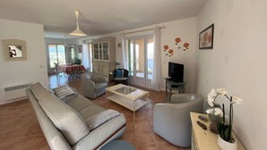 A living room with a sofa, coffee table, and a TV. There is a dining area with a table and chairs in the background. - Superb villa with sea view and private pool (MONTICELLO)