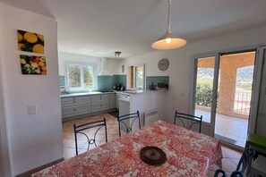 Una cocina con armarios blancos, una mesa de comedor con mantel rojo y blanco, y vistas a un balcón con una montaña al fondo.