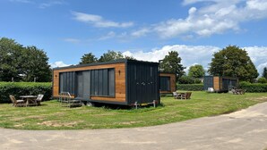 Two modern, compact cabins with large windows, situated on a grassy area with a paved path and outdoor furniture. - Tiny House in Netherlands with Garden (Lathum)