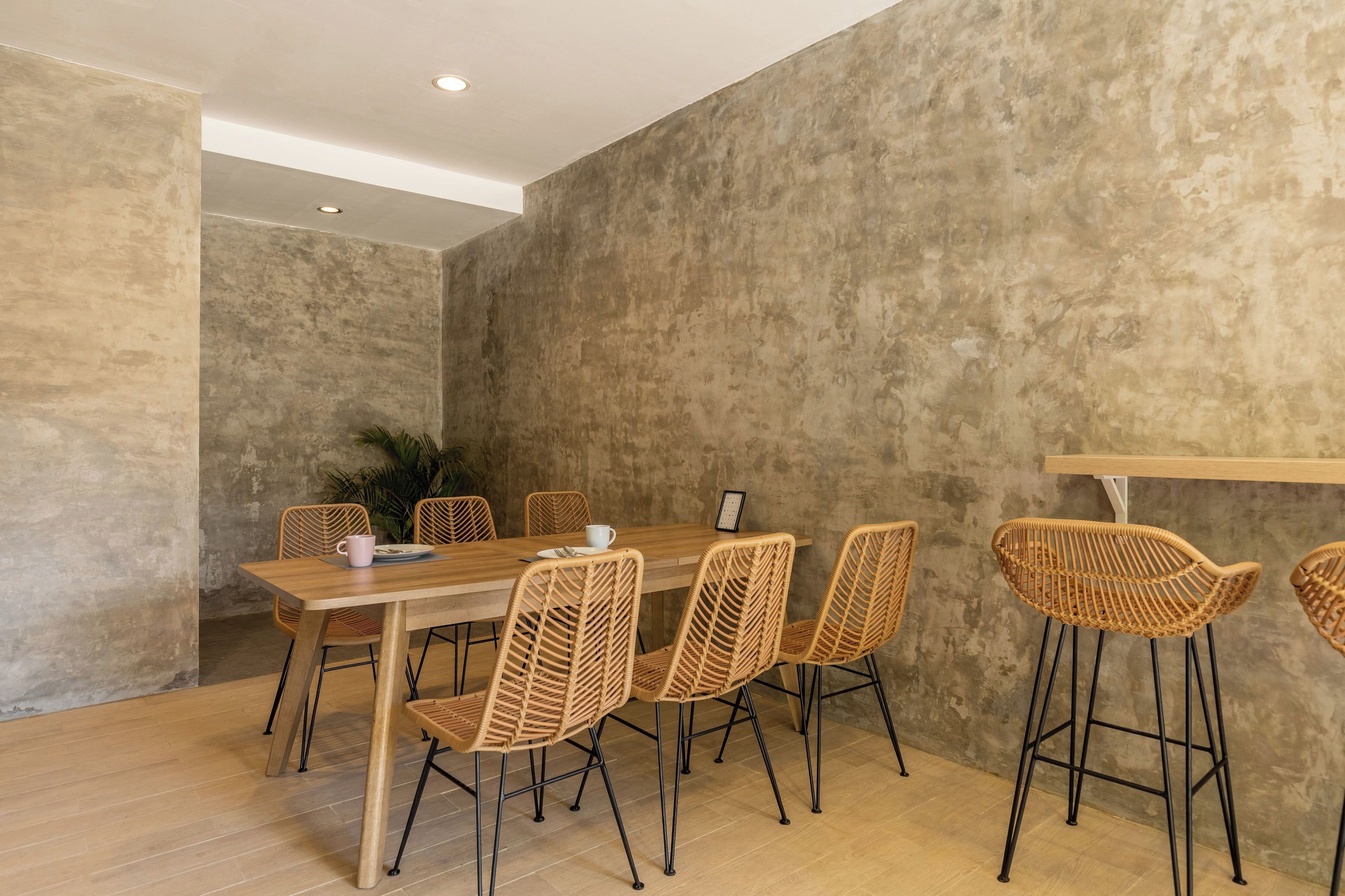 A modern dining area with a wooden table and wicker chairs, a potted plant, and a concrete wall.