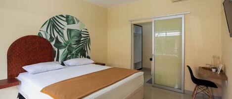 Habitación de hotel con cama, cabecera de madera, puerta corredera, mesita pequeña con una silla y equipo de aire acondicionado.