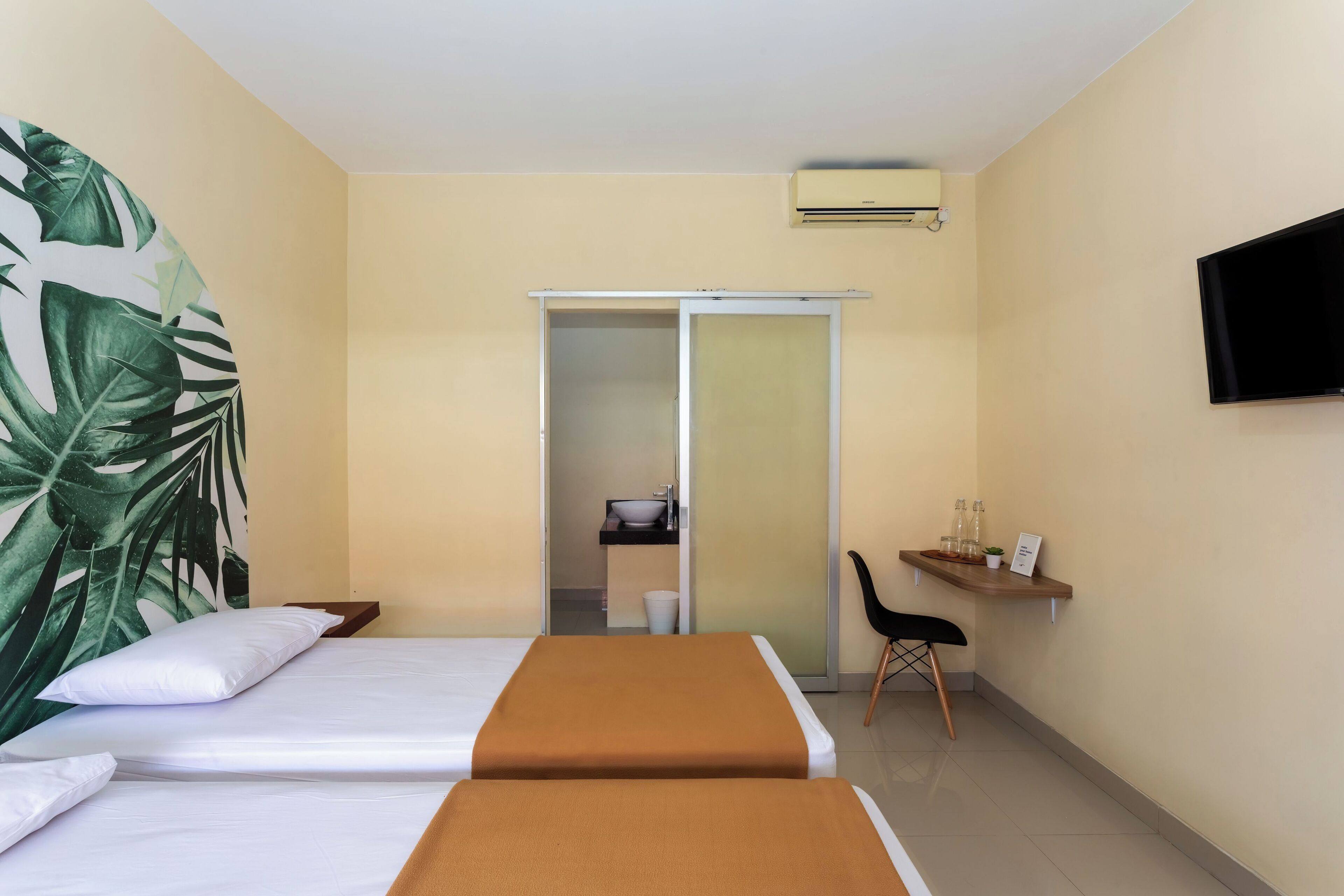 A hotel room with two beds, a TV, a sliding door to a bathroom, and a small shelf with bottles.