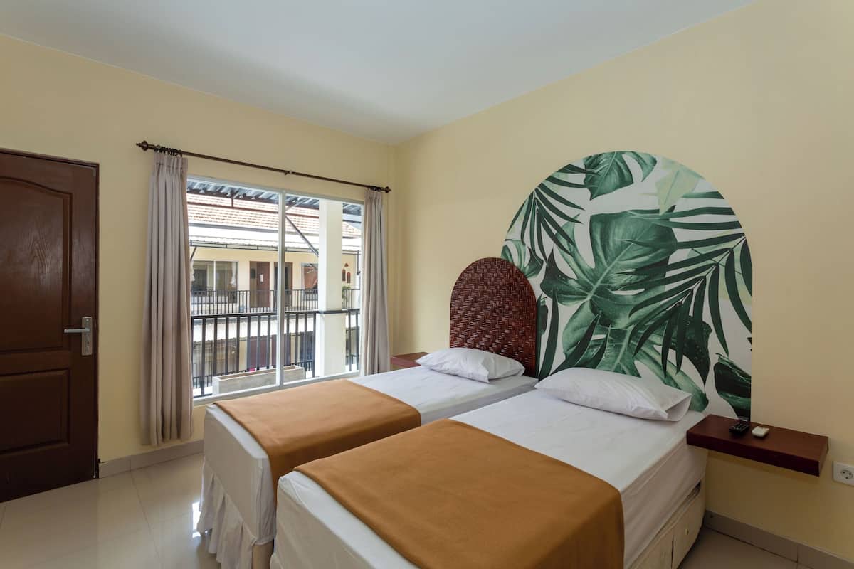 Ein Hotelzimmer mit zwei Betten, einem großen Fenster mit Vorhängen und Blick auf Gebäude sowie einer Wand mit einem tropischen Blattmuster.