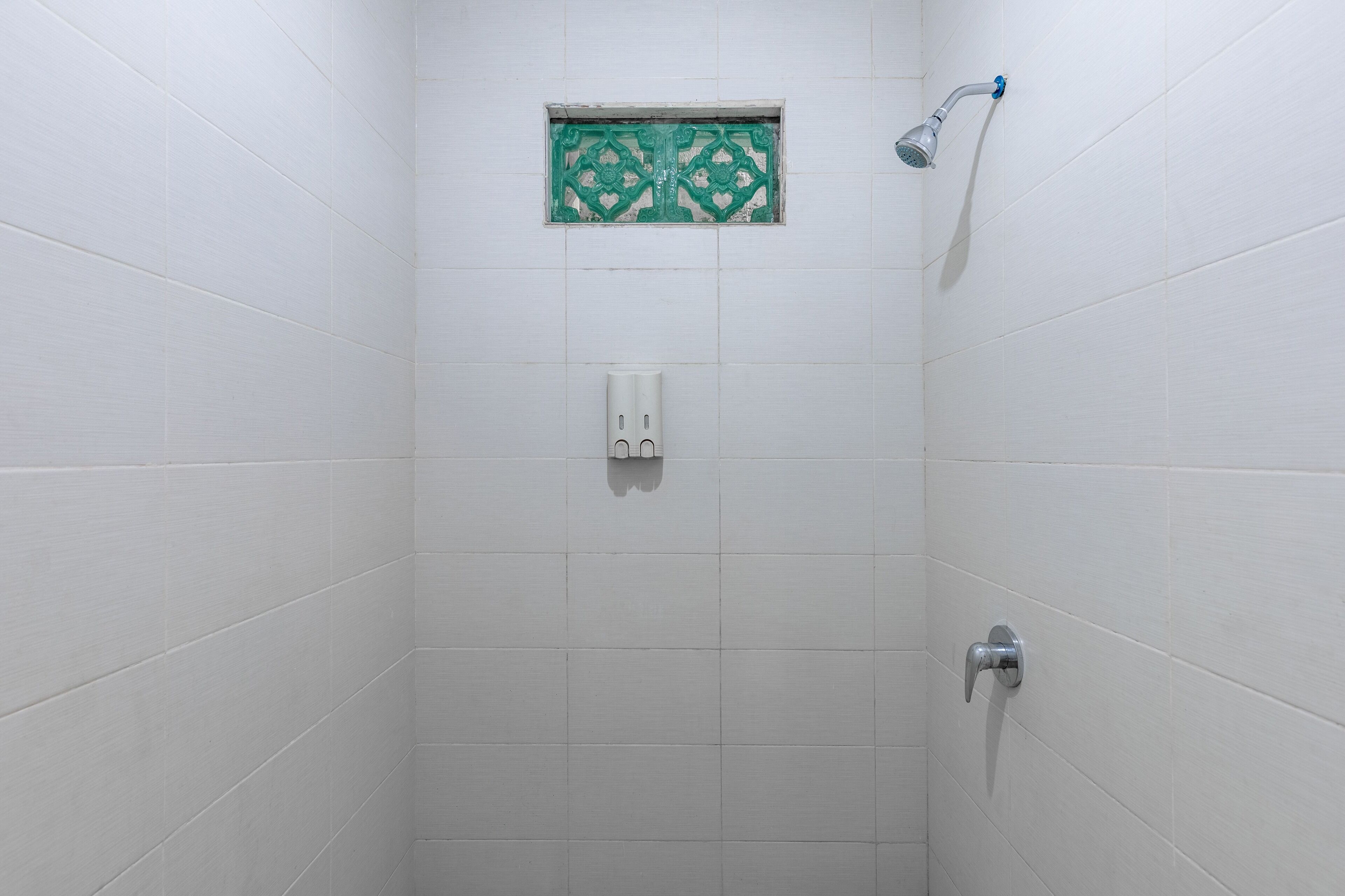 Un área de ducha pequeña y cerrada con azulejos blancos, un azulejo decorativo verde sobre el cabezal de la ducha y un dispensador de jabón blanco.