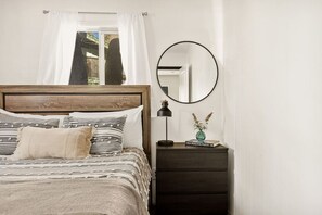 Un dormitorio con cabecera de madera, una cama con ropa de cama a rayas, una mesita de noche con lámpara, un espejo y un pequeño jarrón con flores.