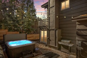 Un jacuzzi en un patio trasero con terraza de madera, una parrilla y luces colgantes.