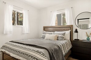 Een slaapkamer met een houten bed, witte gordijnen, een ronde spiegel en een nachtkastje met een lamp.
