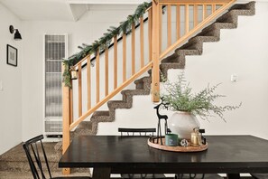 Una escalera con barandas de madera y una guirnalda decorativa, que conduce a un comedor con una mesa dispuesta para dos personas, una planta en maceta y un jarrón.