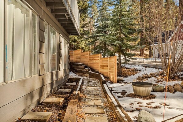 Un sendero conduce a una terraza de madera con un bote de basura metálico y el suelo cubierto de nieve.
