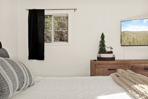Un dormitorio con cama, ventana con vistas a árboles, cómoda con una planta y un cuadro en la pared.