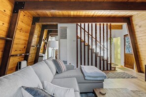 L’intérieur d’un chalet chaleureux, avec un escalier en bois, un canapé d’angle gris et une petite table.