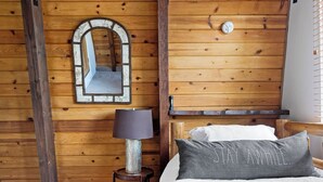 A wooden room with a bed, a lamp, a mirror, and a wall-mounted fire alarm.