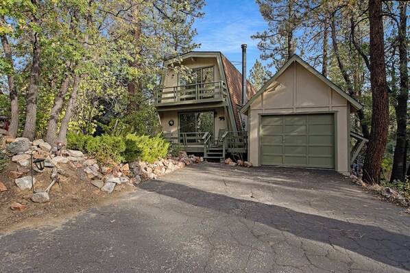 Une cabane à deux étages avec un garage, entourée d’arbres et d’un mur de pierres.