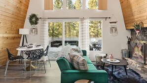 Ein gemütliches Loft mit grünem Sofa, Esstisch und Stühlen, großen Fenstern mit Blick auf Bäume und einem Kamin.