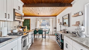 A modern kitchen with white cabinets, a wooden ceiling, and a dining area with a green chair.