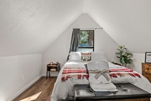 Um quarto com teto inclinado, uma cama com cobertor vermelho e branco, um travesseiro com a inscrição 'RELAX', uma mesinha de cabeceira com relógio e uma janela com cortinas.