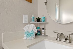 A bathroom with a white sink, a mirror, a shower, and toiletries on a shelf.