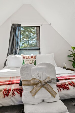 Une chambre à coucher avec un lit, une fenêtre, un oreiller orné du mot « RELAX » et une serviette décorée d’un nœud.