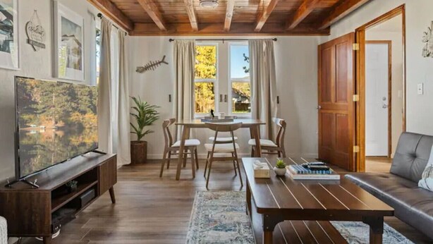 A living room with a wooden ceiling, a TV, a dining table with chairs, a sofa, and a window with curtains.