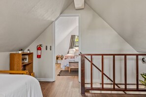 Une chambre avec une bibliothèque en bois, un extincteur, un lit recouvert d’une couette blanche et un escalier en bois.
