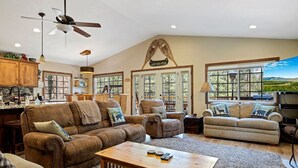 A spacious living room with a large brown sofa, two armchairs, and a coffee table. There are large windows with a view of the outdoors.