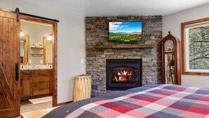 A bedroom with a stone fireplace, a television mounted above it, and a large window with a view of trees.