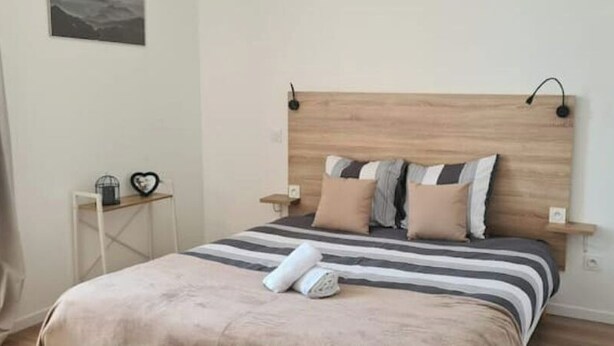 Una cama bien hecha con ropa de cama a rayas y varios almohadones, una mesita de noche con una lámpara, y un pequeño estante con objetos decorativos.