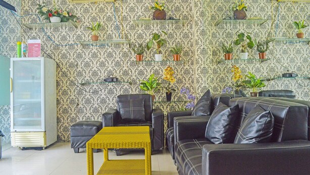 A living room with a sofa, chairs, a small table, and a refrigerator. There are shelves with plants on the wall.