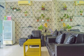 Een woonkamer met een bank, stoelen, een tafeltje en een koelkast. Er staan planten op een wandplank.