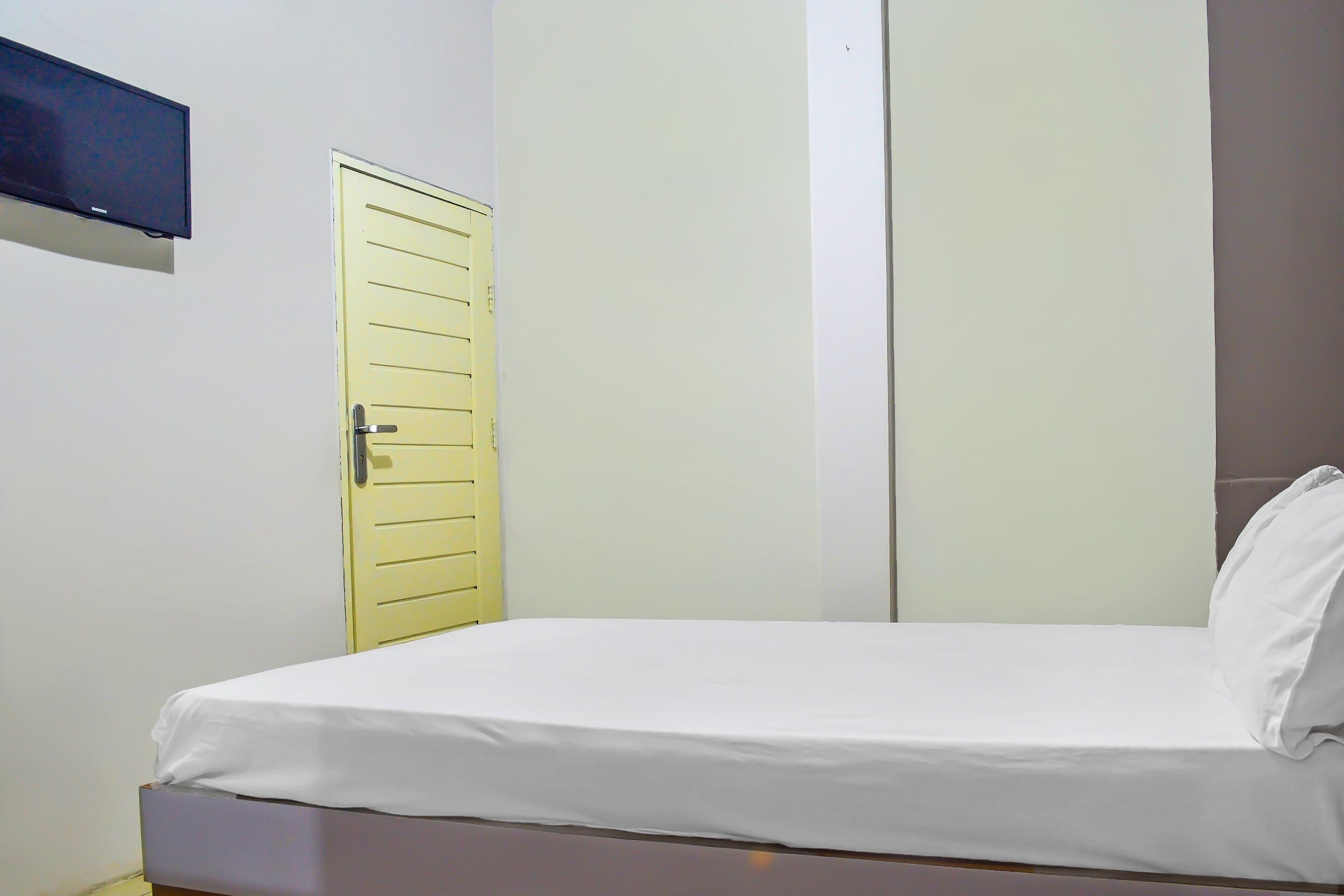 A single bed with a white mattress and pillow, a yellow door, and a wall-mounted television.