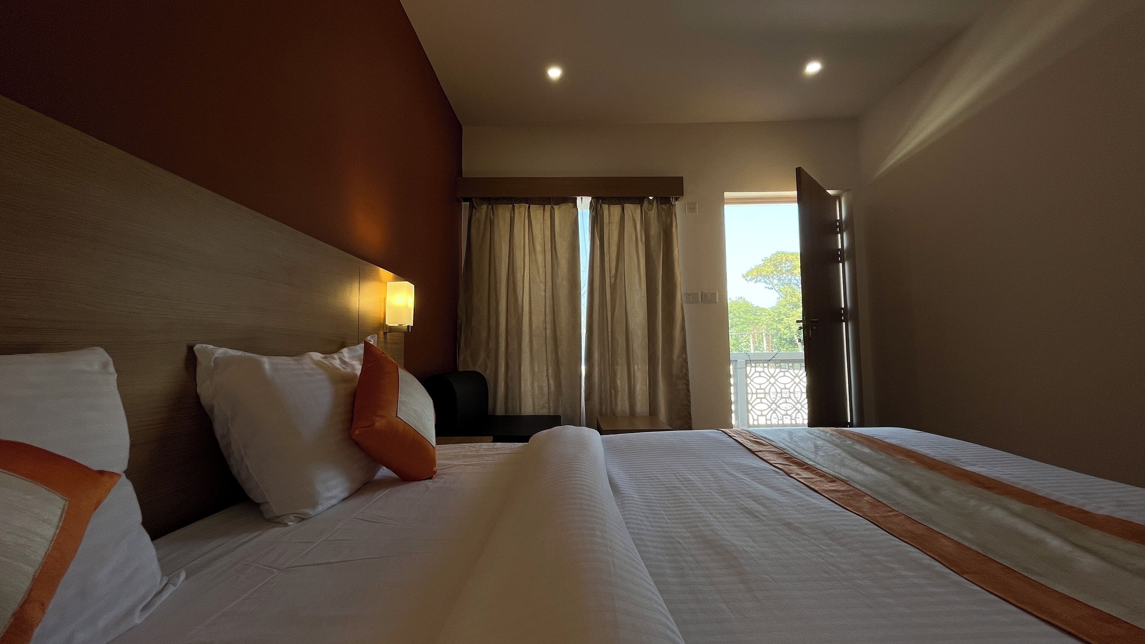 A hotel room with a large bed, a bedside table, a lamp, and a view of the outdoors through a window with curtains.