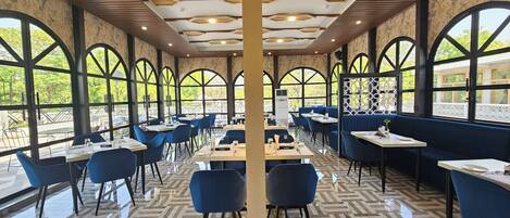 A restaurant with a modern interior, featuring a herringbone patterned floor, blue seating, and large arched windows offering views of the outdoors.