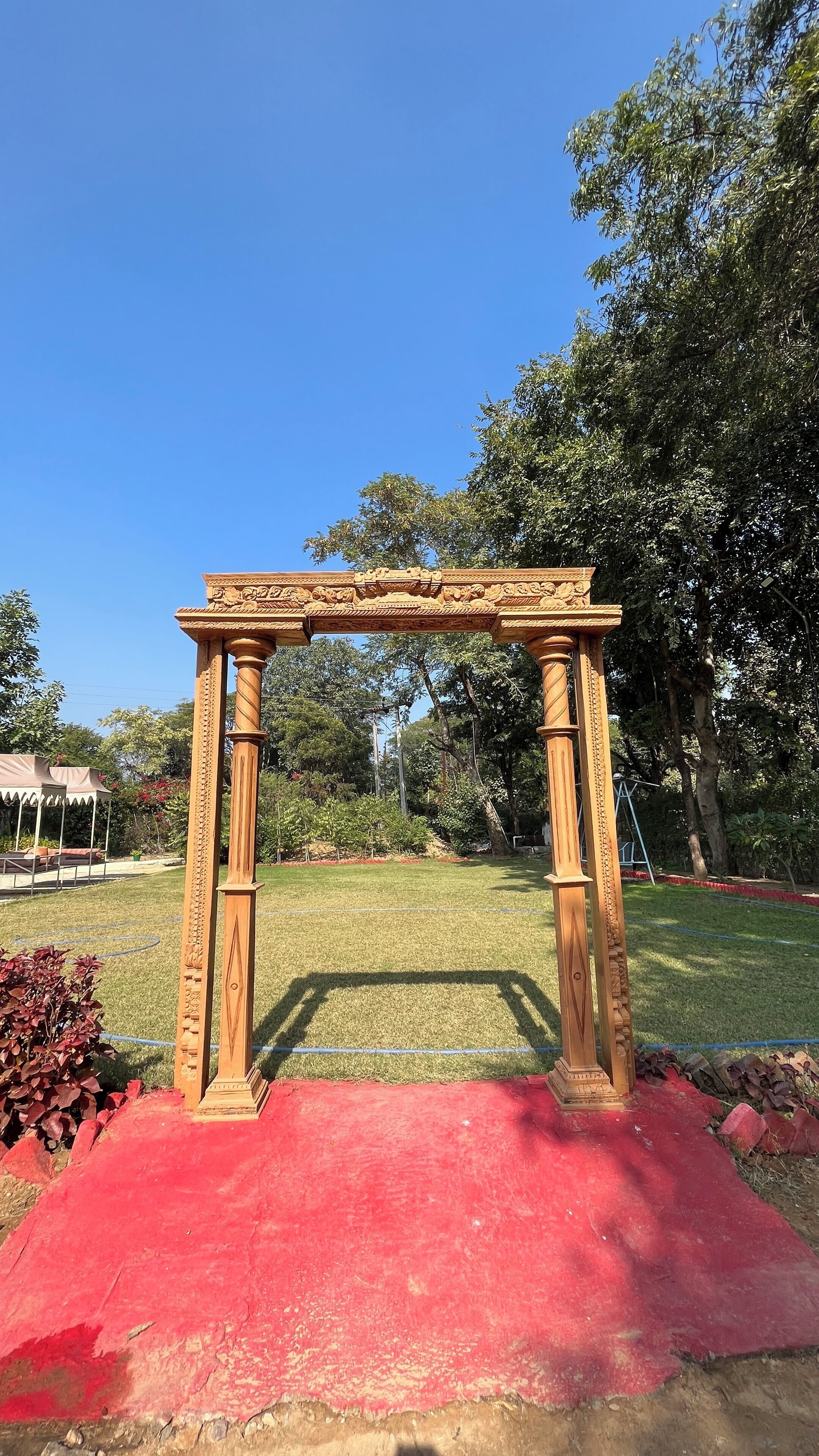 Un arco de madera con intrincados grabados, sobre una alfombra roja en un entorno similar a un parque, con árboles y un cielo azul despejado.