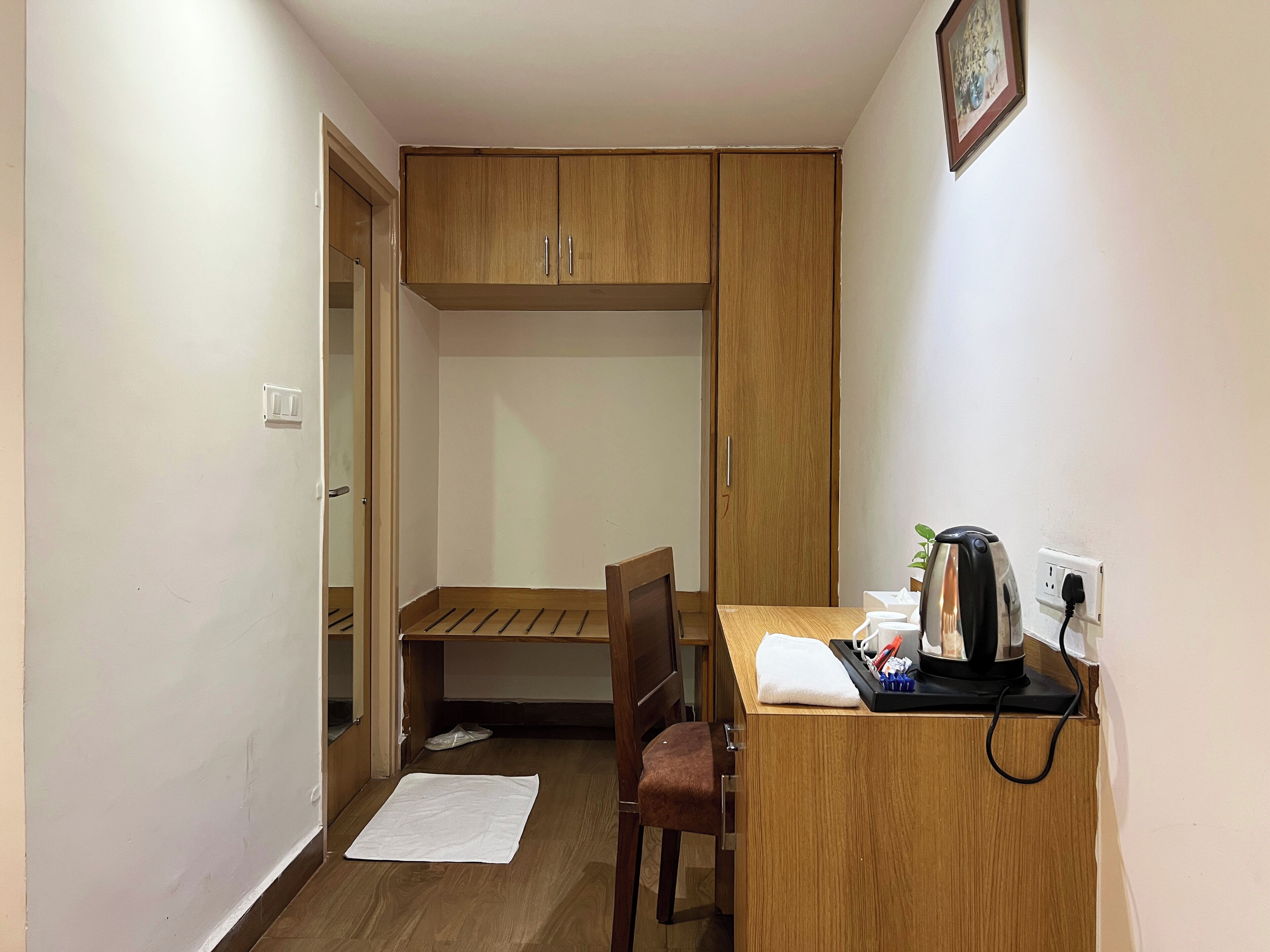 Una habitación pequeña con un escritorio de madera, una silla y una cama con cabecera de madera. Hay una toalla blanca en el suelo y una mesita con una tetera y tazas.