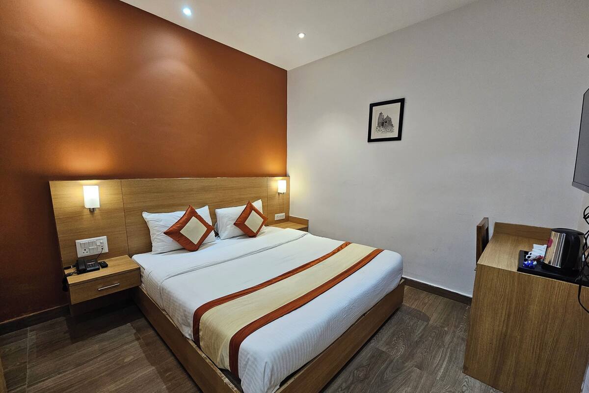 A hotel room with a large bed, wooden headboard, bedside lamps, a nightstand, a desk with a chair, and a framed picture on the wall.
