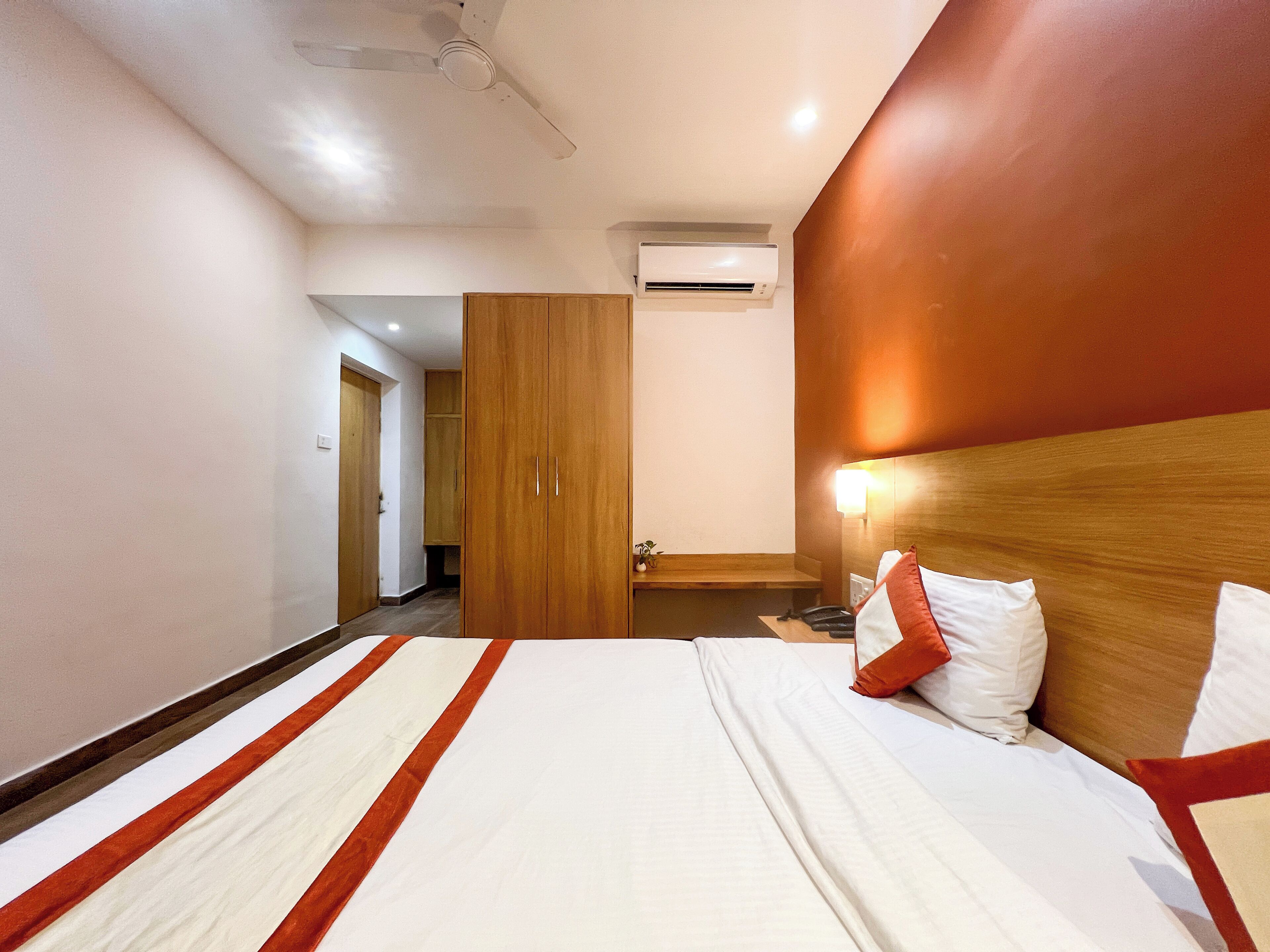 A hotel room with a bed, a wooden wardrobe, an air conditioning unit, and a bedside table with a lamp.