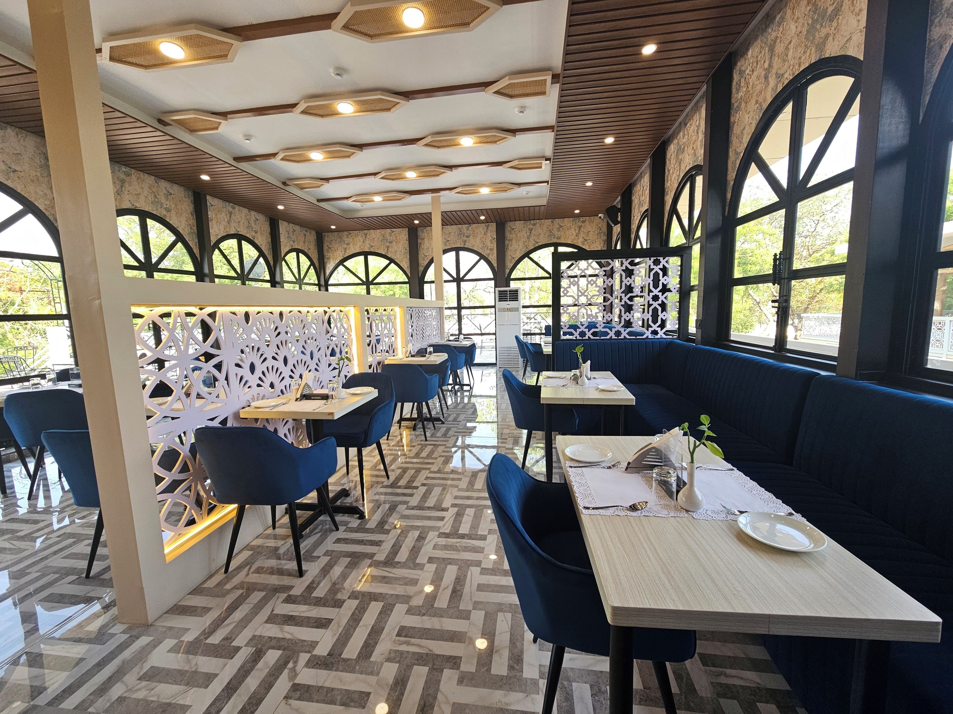 Un restaurante con una decoración moderna, que presenta un suelo con diseño de espiga, asientos tapizados en azul y grandes ventanales que dejan entrar luz natural.